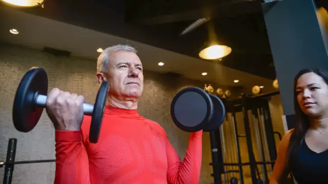 Homem mais velho treinando com halteres ao lado de uma treinadora