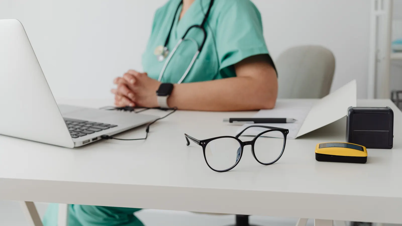 Mesa de consultório com óculos, notebook e profissional de saúde ao fundo