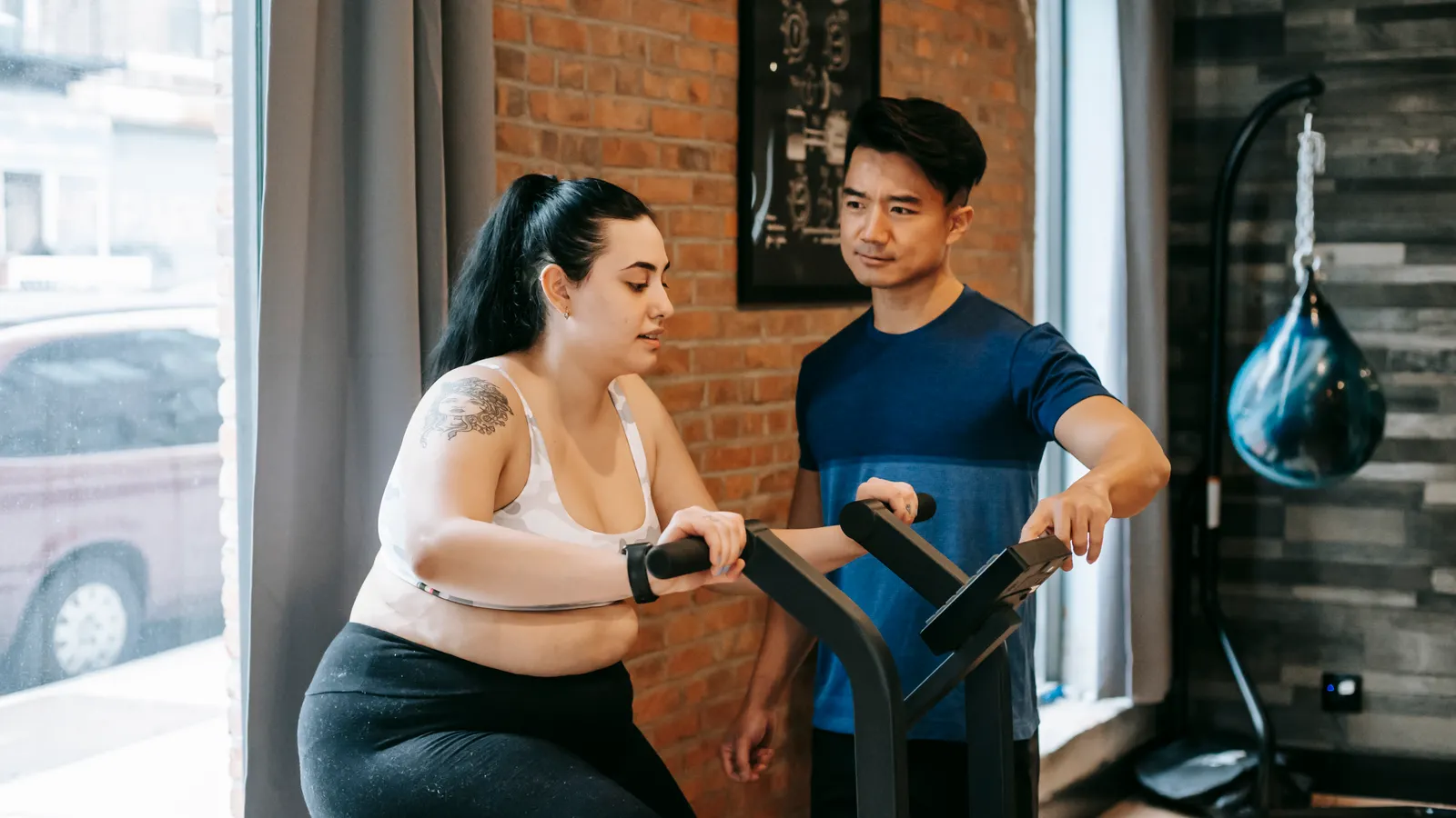 Personal trainer orientando uma aluna que pedala numa bicicleta ergométrica, numa academia com parede de tijolos